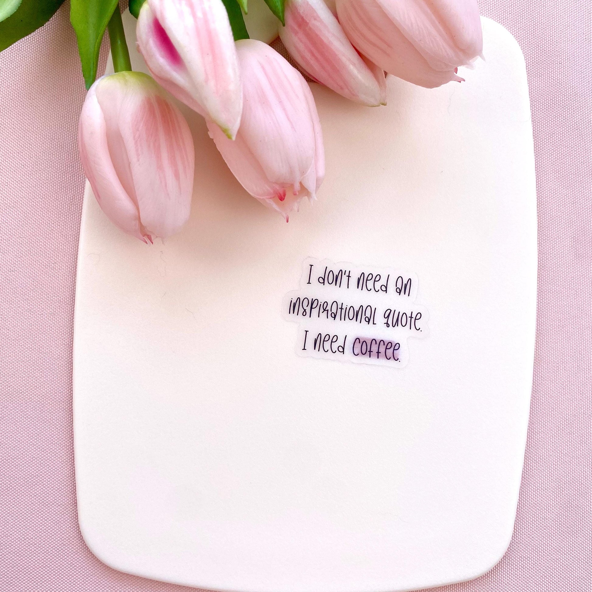 Clear vinyl sticker with black text 'I don't need an inspirational quote, I need coffee' shown on a white background with pink tulips