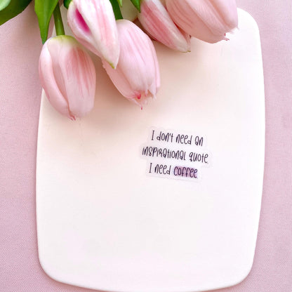 Clear vinyl sticker with black text 'I don't need an inspirational quote, I need coffee' shown on a white background with pink tulips