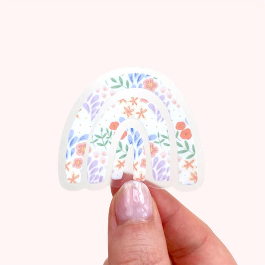 Decorative transparent rainbow sticker with floral design held by a hand on a white background
