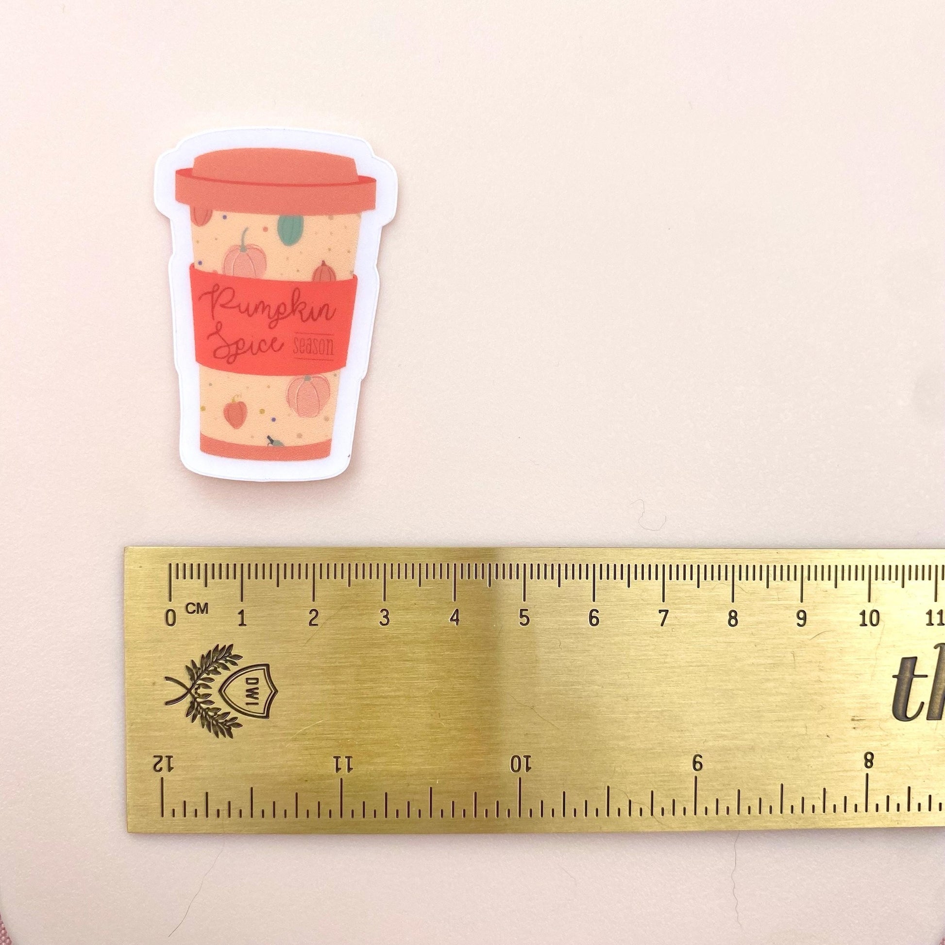 Sticker of a pumpkin spice latte cup next to a ruler on a beige background