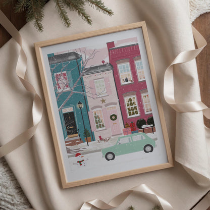 Framed illustration of a colourful winter street scene with houses and a car in the snow, surrounded by ribbons on a soft surface.