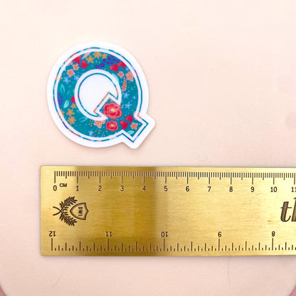Decorative letter 'Q' sticker with floral design next to a ruler on a beige background