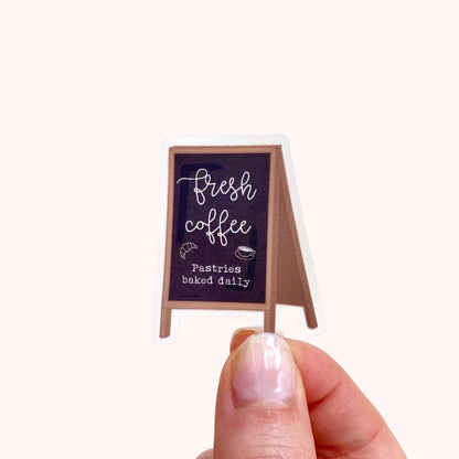 Sticker with a chalkboard sign held by a hand with 'Fresh coffee' and 'Pastries baked daily' text on a white background.