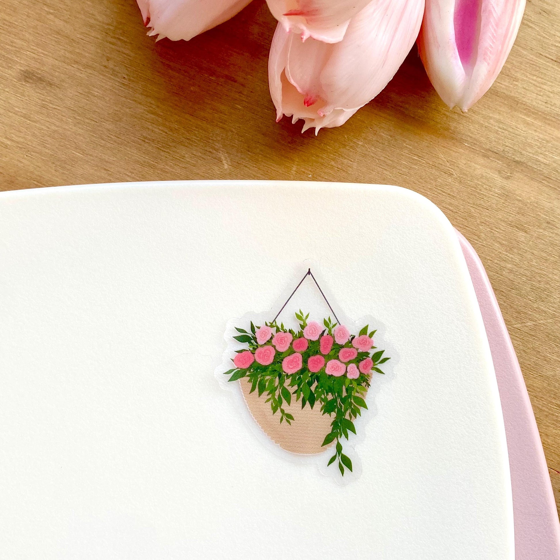 A sticker with a hanging planter with pink flowers against a white surface, with pink tulips in the background.