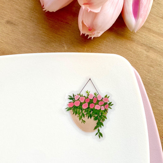 A sticker with a hanging planter with pink flowers against a white surface, with pink tulips in the background.