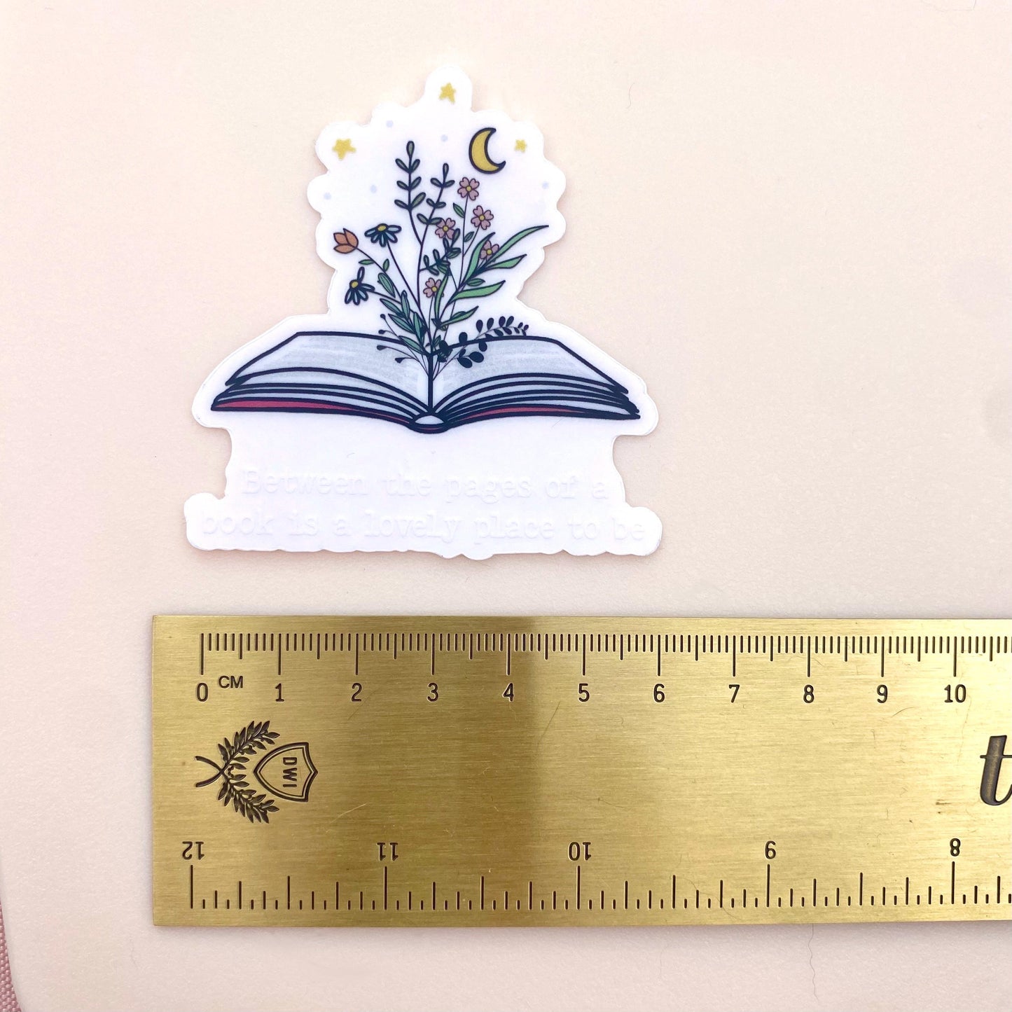 Sticker of an open book with floral design next to a ruler for scale on a beige background