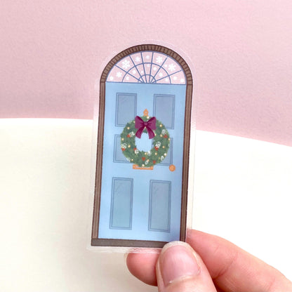 A sticker with a blue door decorated for Christmas being held by hand over a pink and cream background.