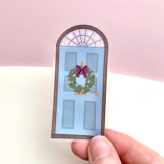 A sticker with a blue door decorated for Christmas being held by hand over a pink and cream background.