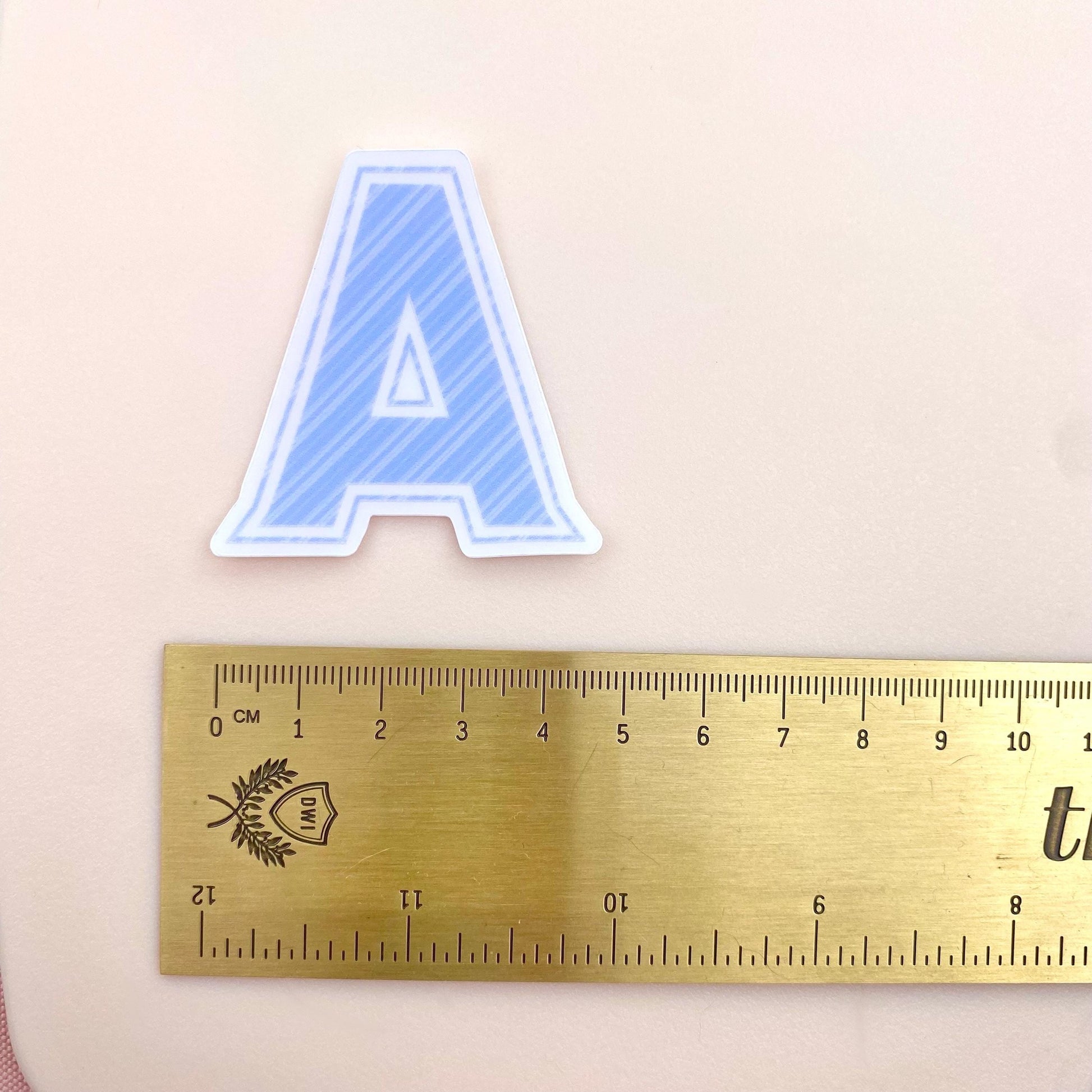 Blue striped letter 'A' sticker next to a gold ruler for scale on a beige background