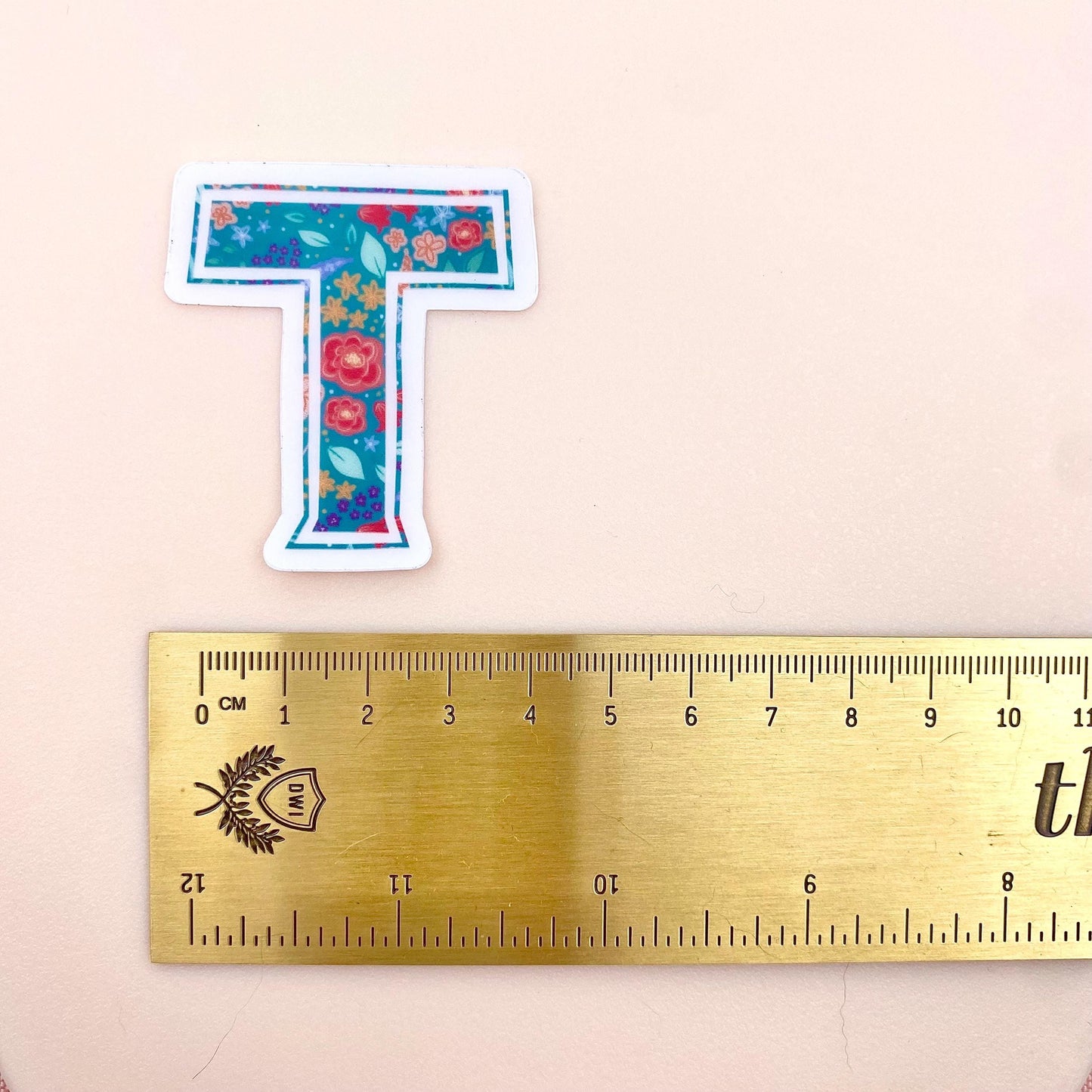 Floral letter 'T' sticker next to a ruler on a beige background