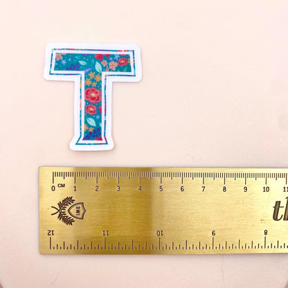 Floral letter 'T' sticker next to a ruler on a beige background