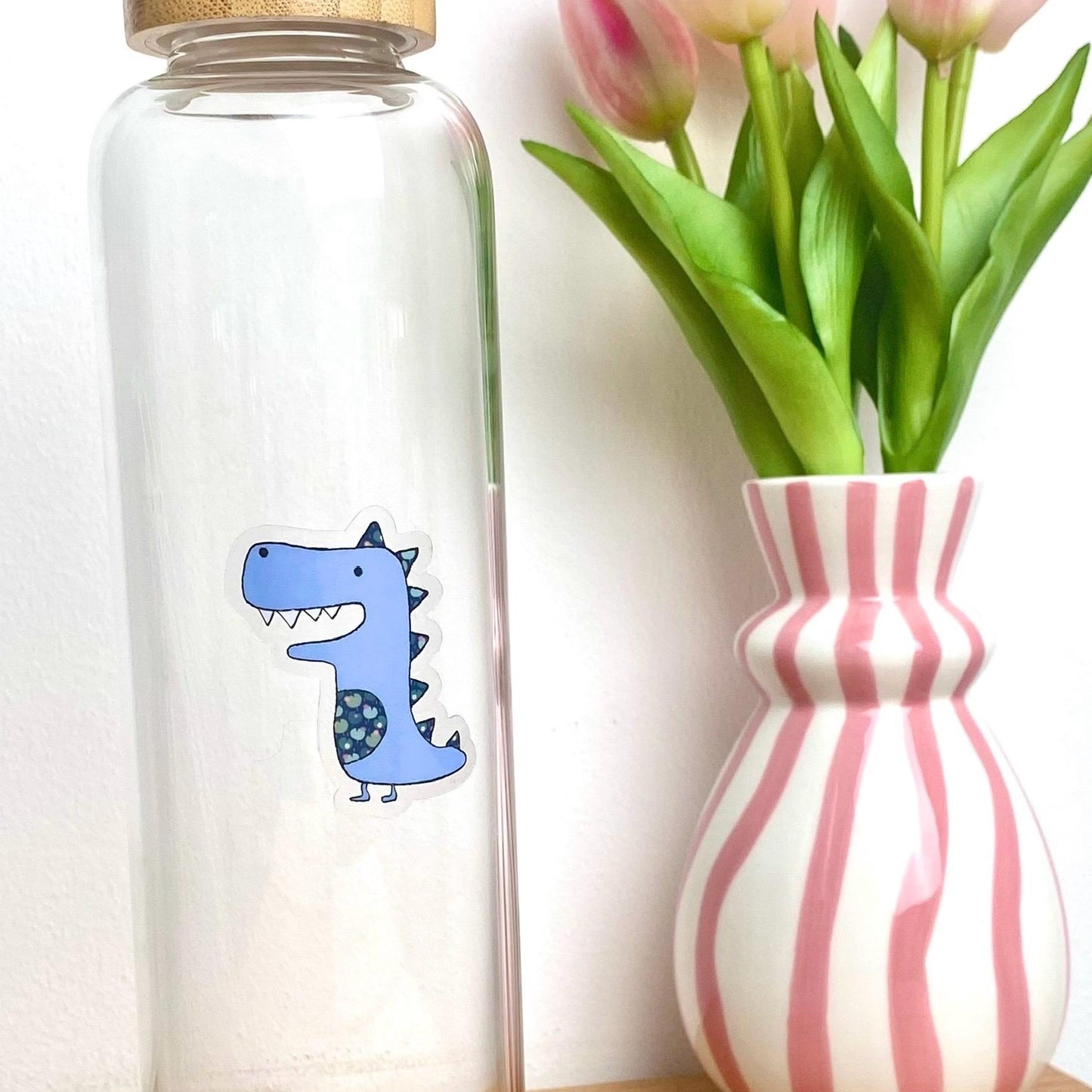 Clear glass bottle with a blue dinosaur sticker next to a striped vase with tulips