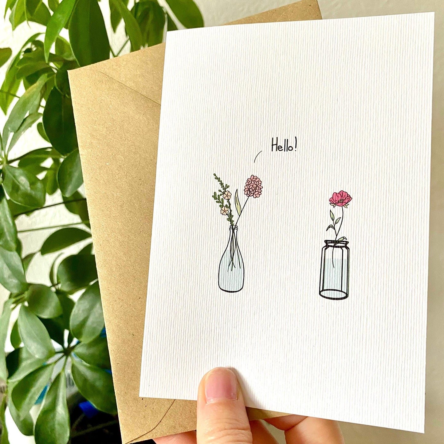 Greeting card with floral illustrations and 'Hello' text, held in front of a plant.