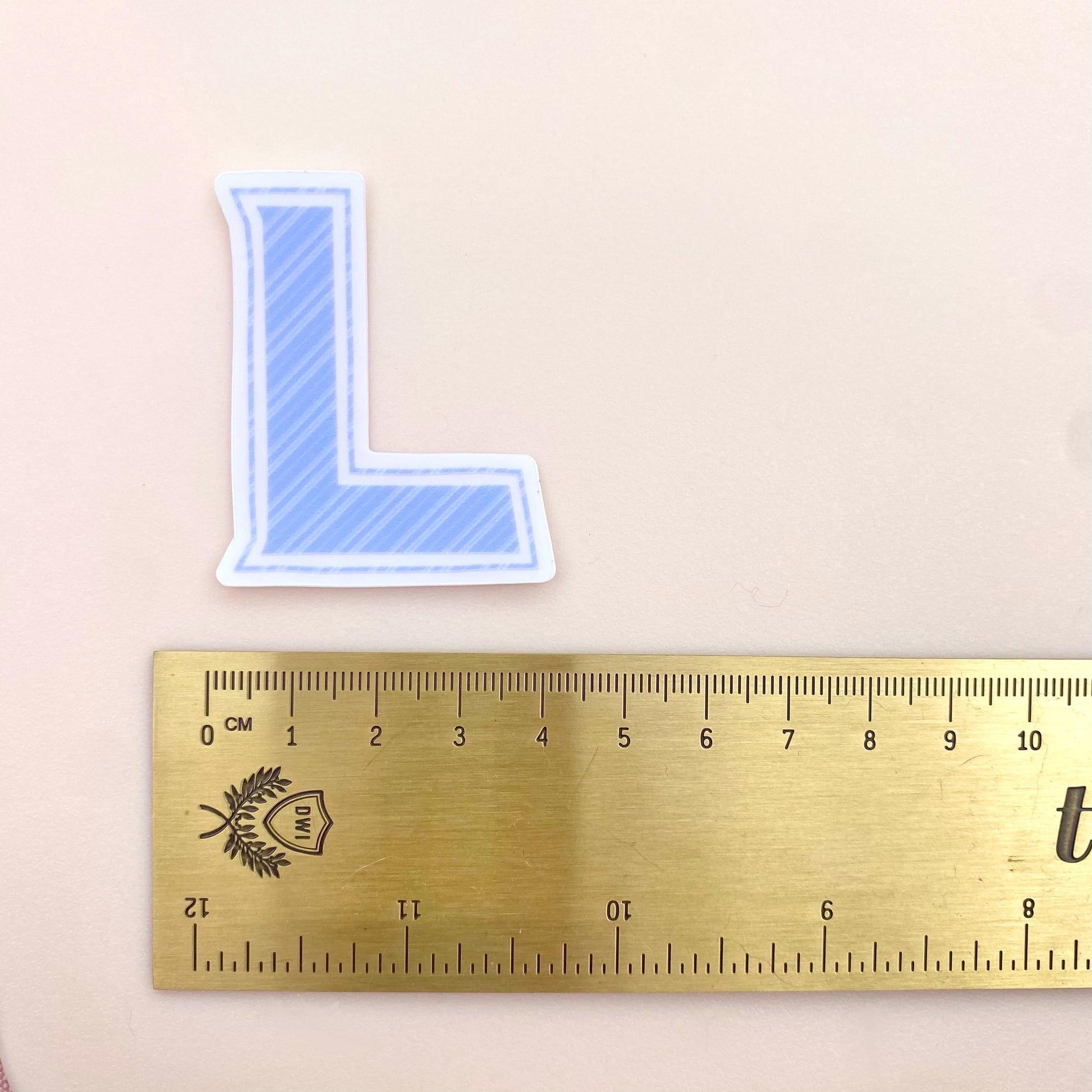 Blue striped letter 'L' sticker next to a ruler on a beige background