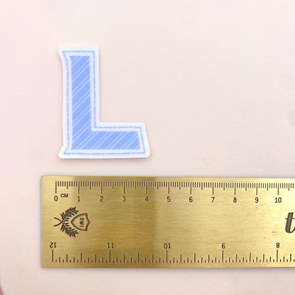 Blue striped letter 'L' sticker next to a ruler on a beige background