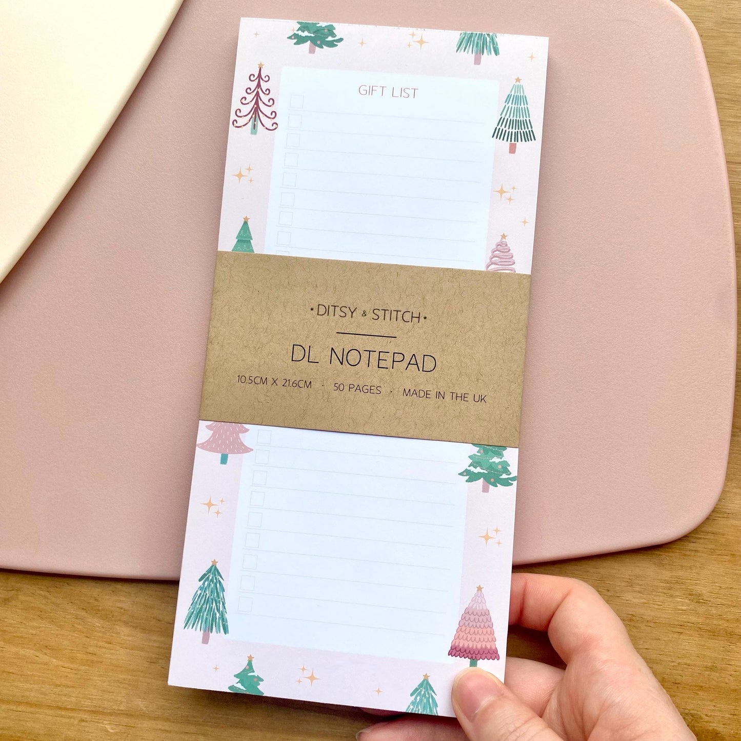 Slim pink gift list notepad with Christmas tree design held by a hand on a wooden surface.