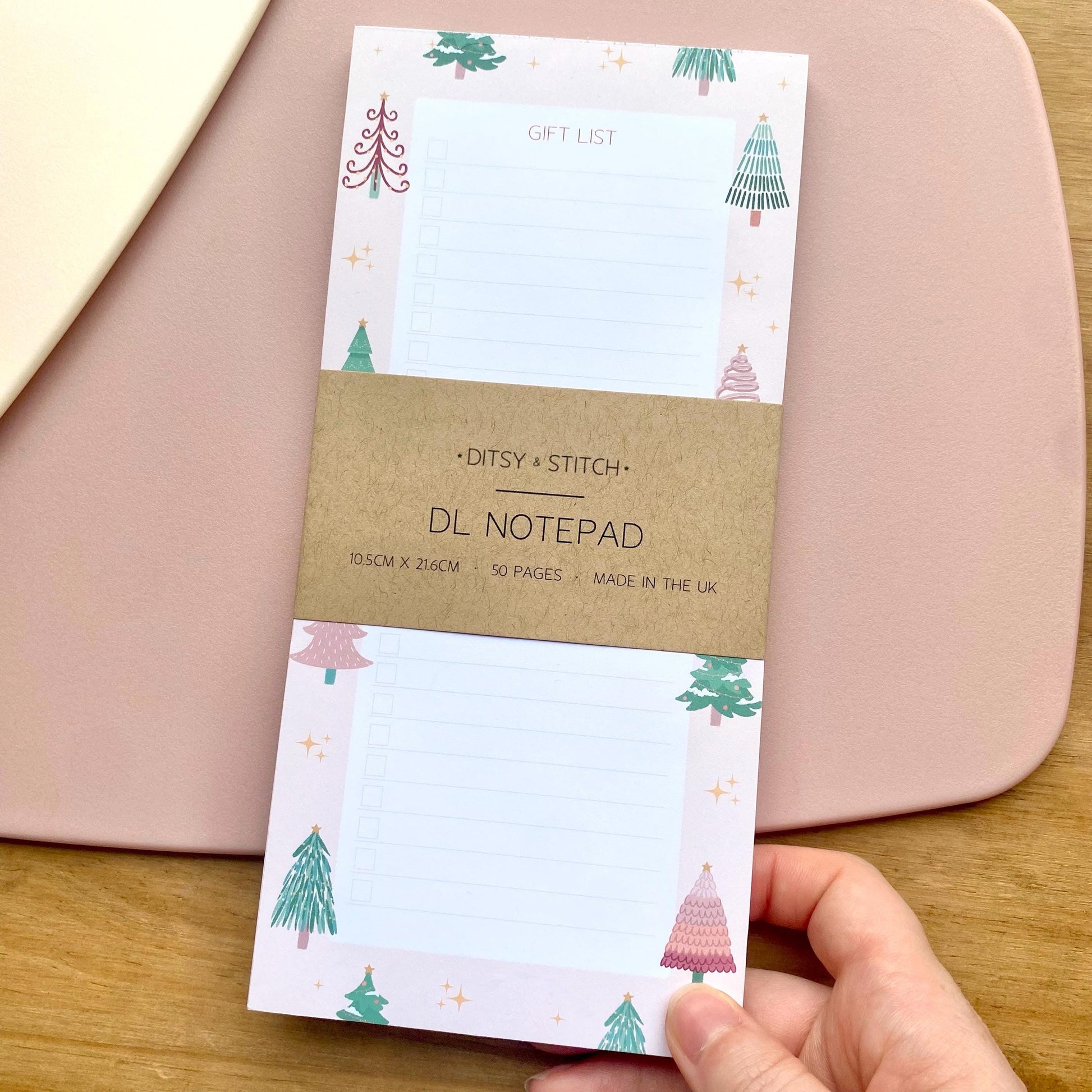 Slim pink gift list notepad with Christmas tree design held by a hand on a wooden surface.