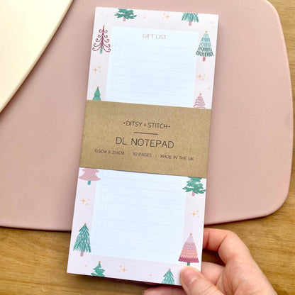 Slim pink gift list notepad with Christmas tree design held by a hand on a wooden surface.