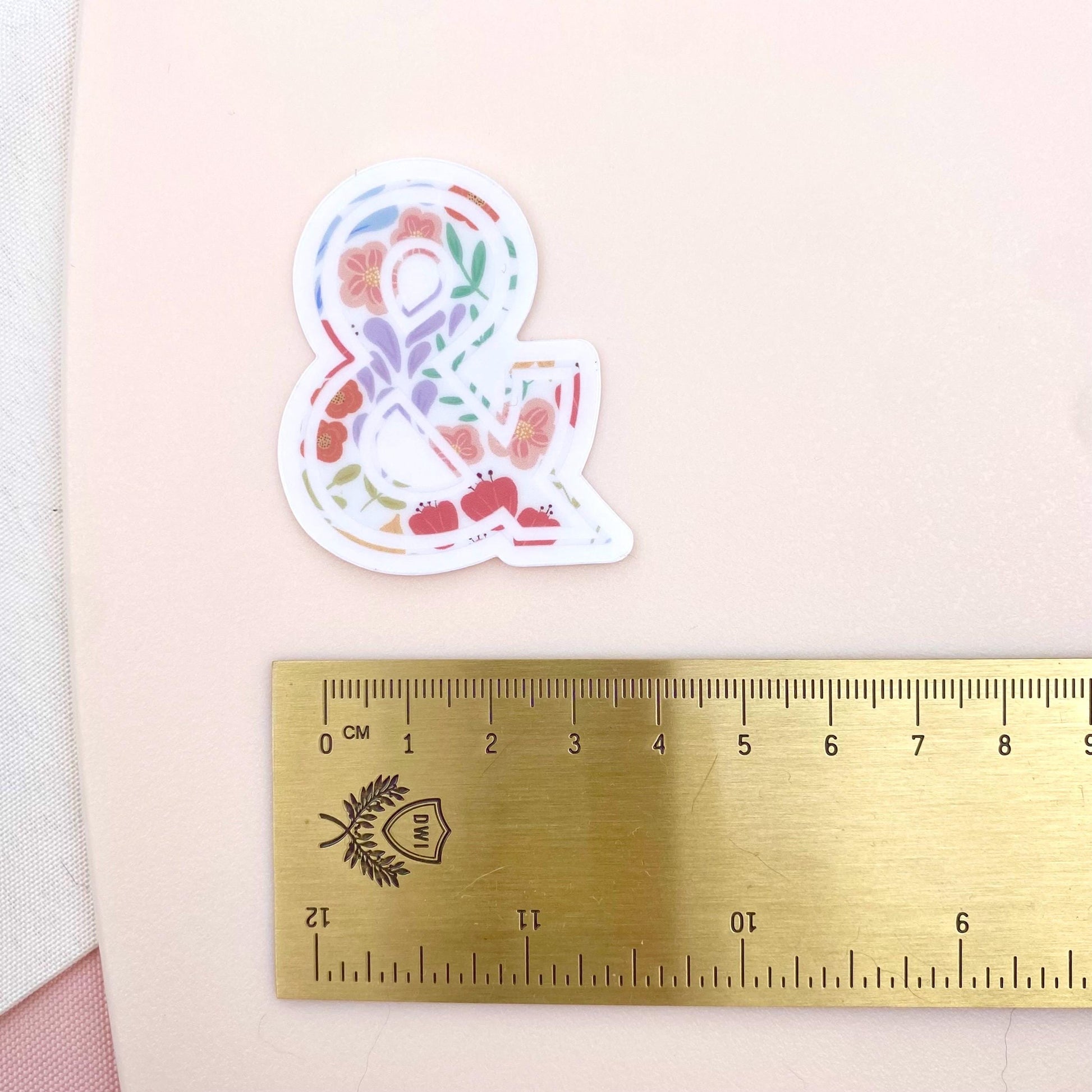 Decorative ampersand sticker with floral pattern next to a ruler on a pink background