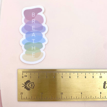 Colorful 'BREATHE' sticker with a gold ruler for scale on a light pink background