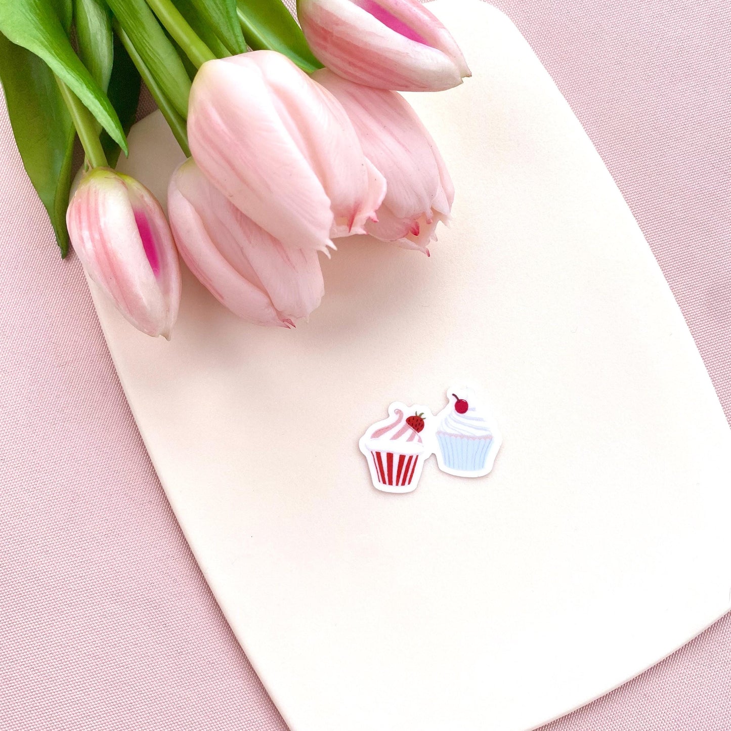 A sticker with two tiny cupcakes in pink and blue against a cream background with pink tulips.