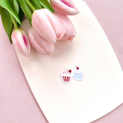 A sticker with two tiny cupcakes in pink and blue against a cream background with pink tulips.