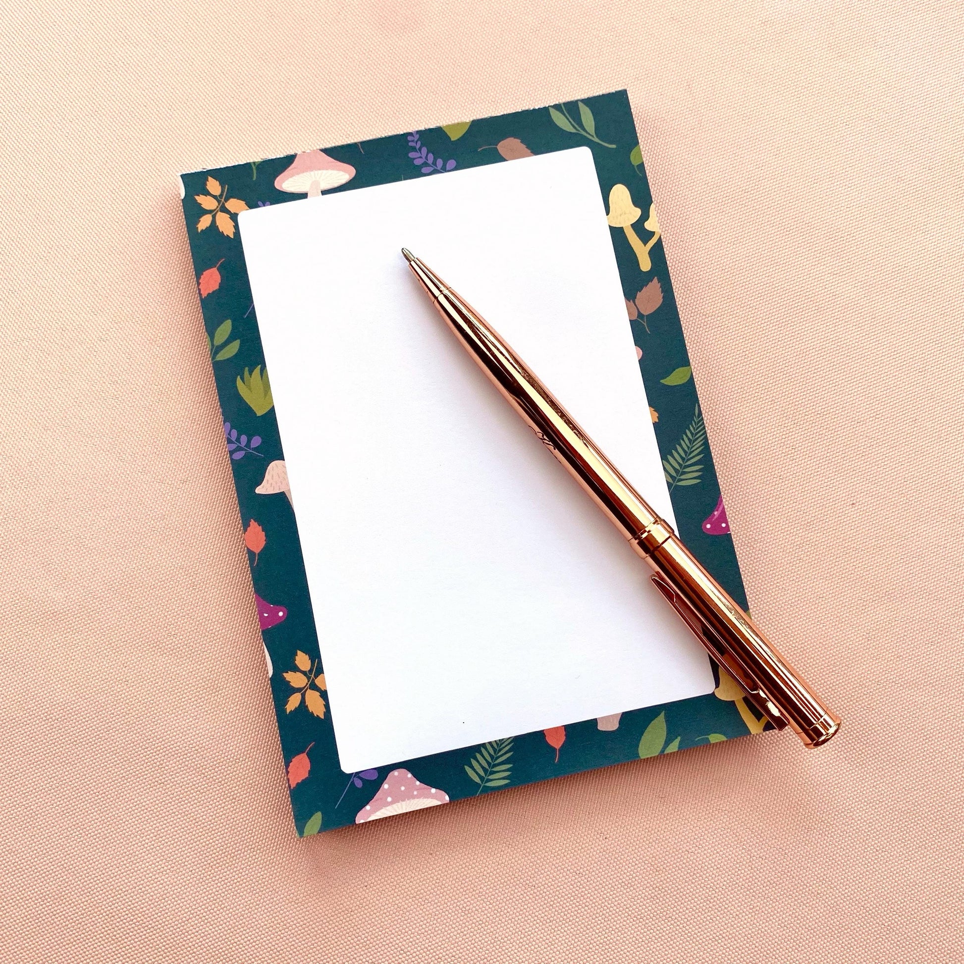 Notepad with mushroom and leaf patterned border on a white background with a rose gold pen