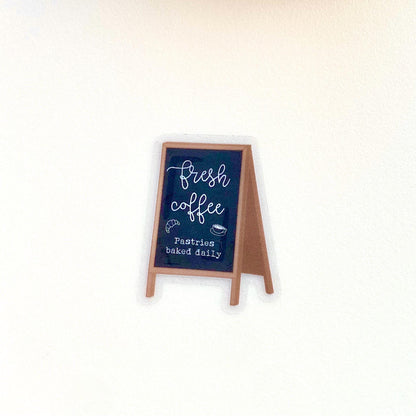 Sticker with a chalkboard sign held by a hand with 'Fresh coffee' and 'Pastries baked daily' text on a white background.