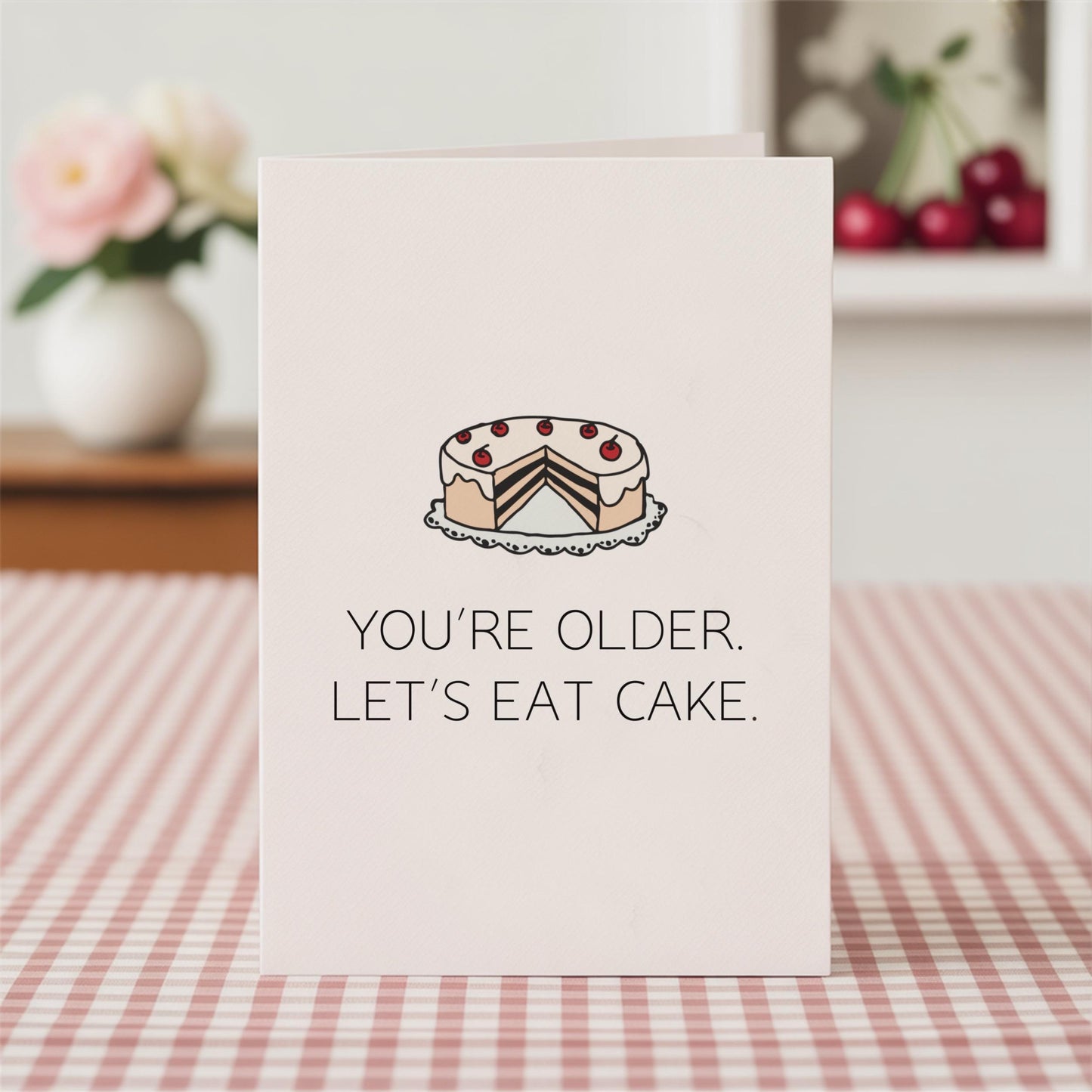 Card with a cake illustration and text 'You're older. Let's eat cake.' on a checkered tablecloth.