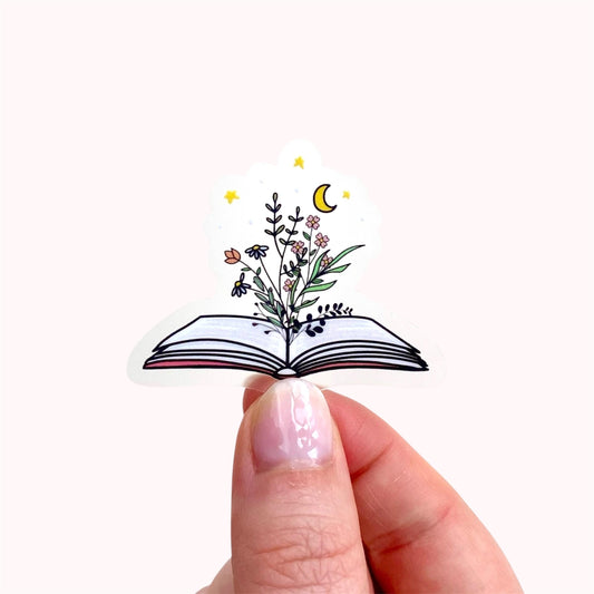 Decorative sticker of an open book with floral designs held between fingers on a white background