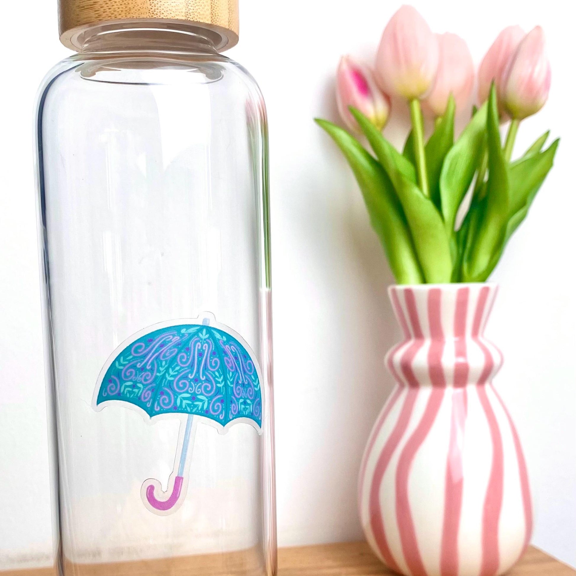 Clear glass water bottle with a decorative umbrella sticker next to a striped vase with tulips.