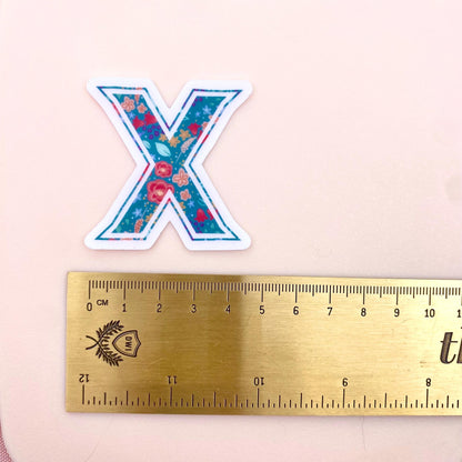 Floral-patterned letter 'X' sticker next to a ruler on a pink background