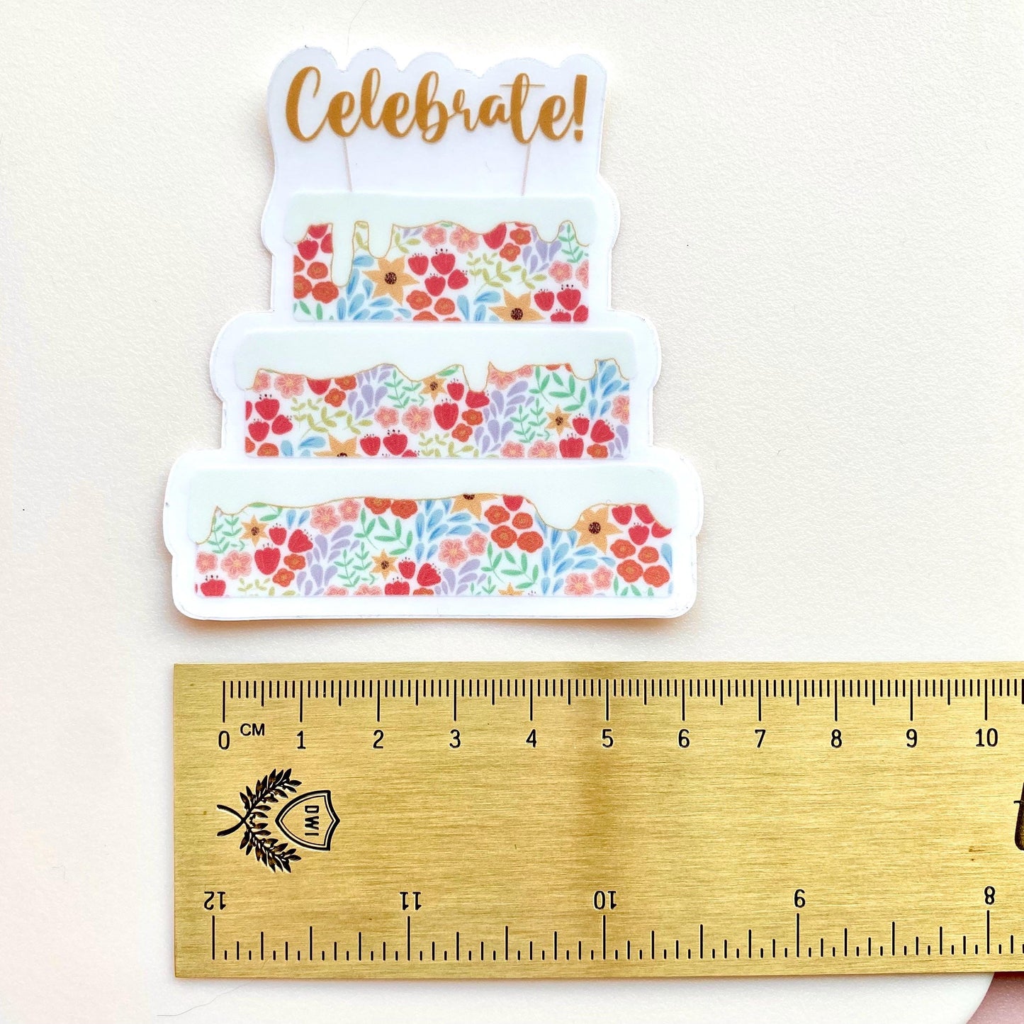 Decorative cake sticker with floral patterns and 'Celebrate!' text, shown with a wooden ruler for scale.