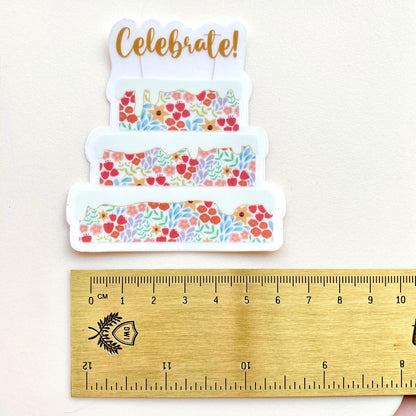 Decorative cake sticker with floral patterns and 'Celebrate!' text, shown with a wooden ruler for scale.