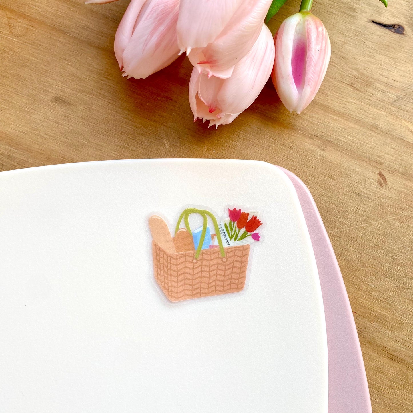 Sticker of a picnic basket with flowers on a white surface, with pink tulips in the background.