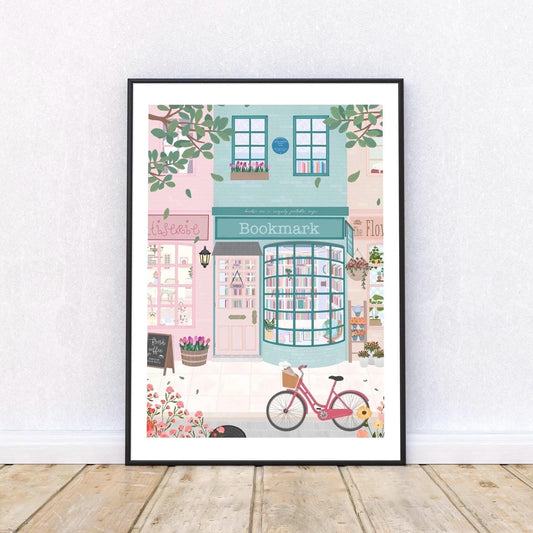 Framed artwork of a quaint street scene with a bookstore, pink bicycle, and greenery.