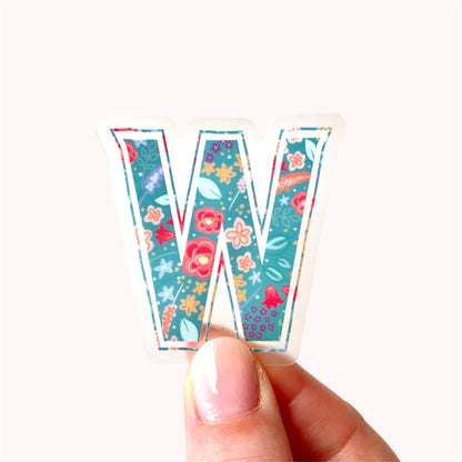 Hand holding a colourful letter 'W' sticker with floral patterns on a white background