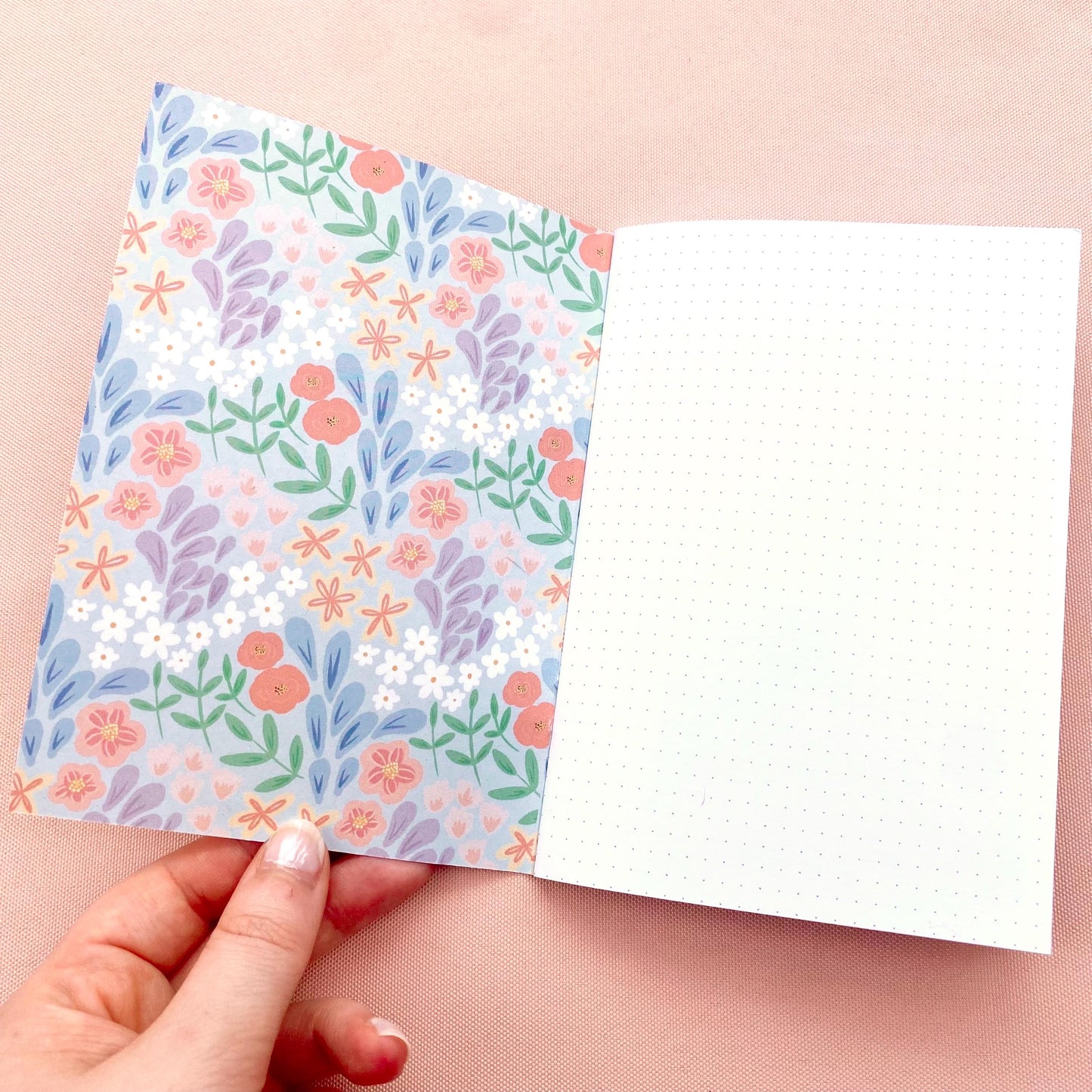Notebook with dotted white pages and floral pattern on the inner cover held open by a hand on a pink background