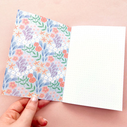 Notebook with dotted white pages and floral pattern on the inner cover held open by a hand on a pink background