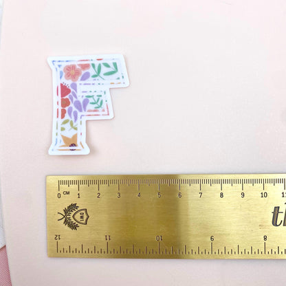 Floral letter 'F' sticker next to a ruler on a pink background