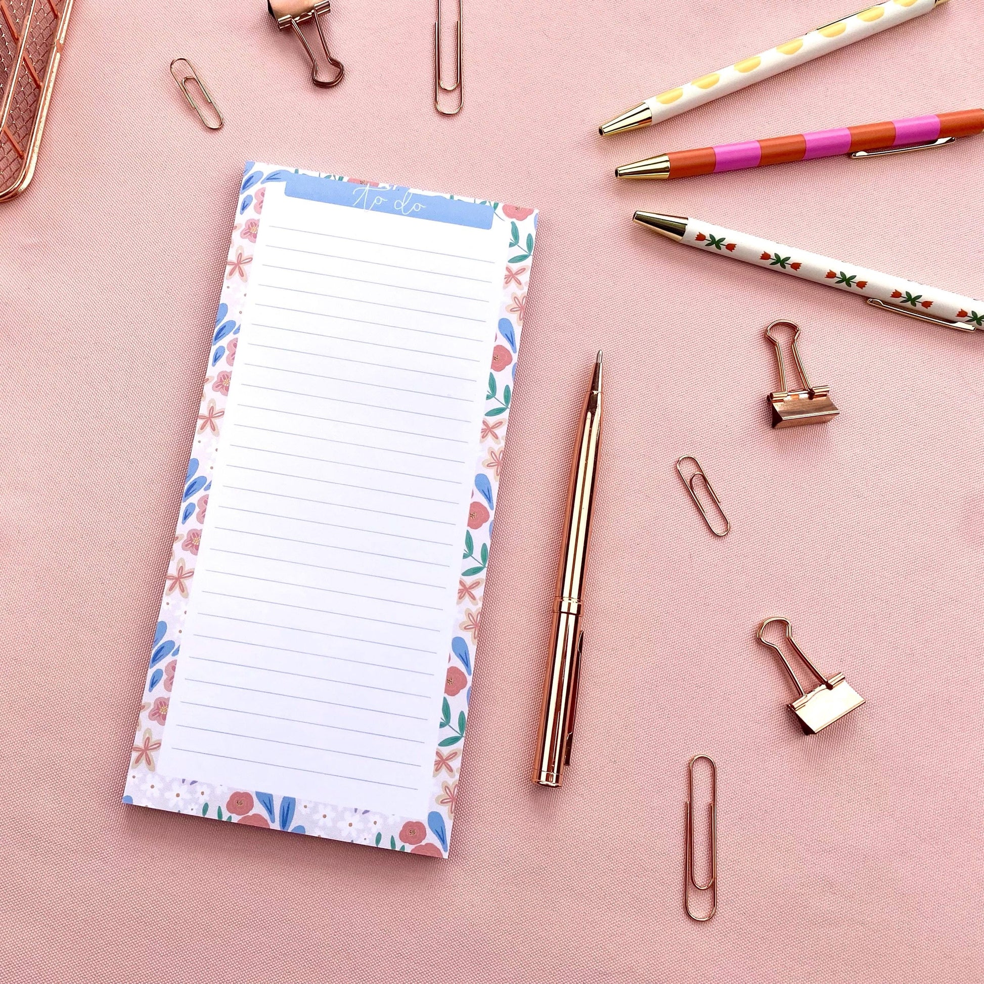 Notepad with floral border, pen, and paper clips on a pink background