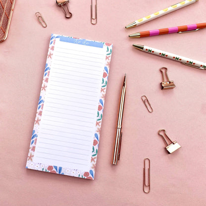 Notepad with floral border, pen, and paper clips on a pink background