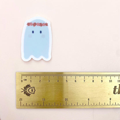 Cartoon ghost sticker with a flower crown with a gold ruler for scale on a light pink background