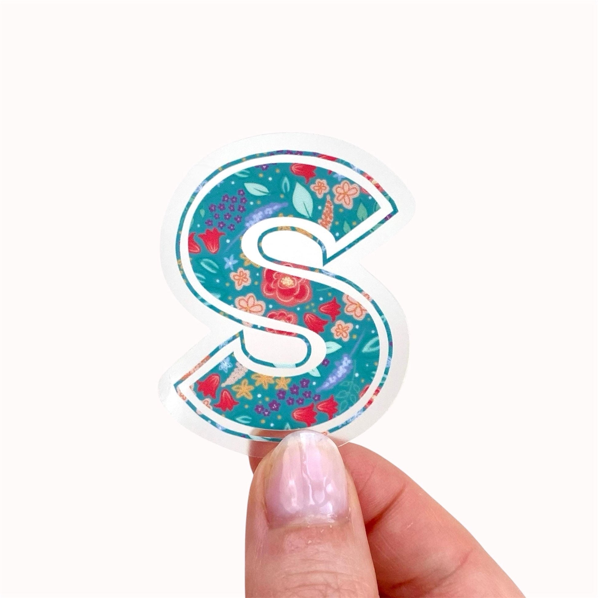 Decorative letter 'S' sticker with floral patterns held by a hand on a white background