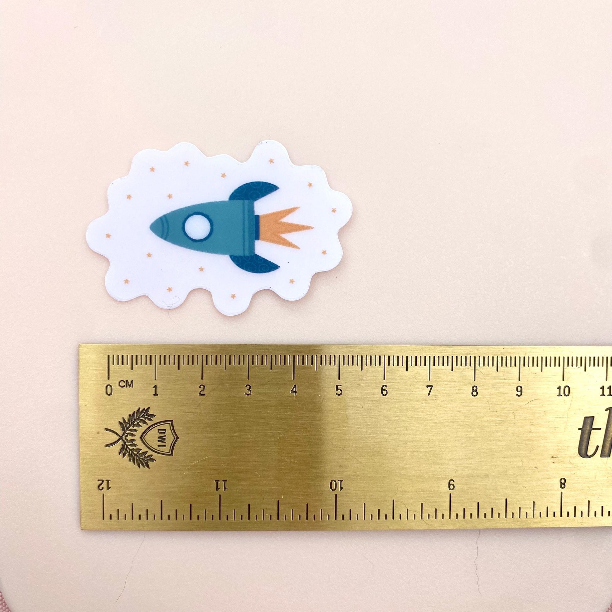 Transparent sticker with teal blue rocket against a cream background with a gold ruler for scale.