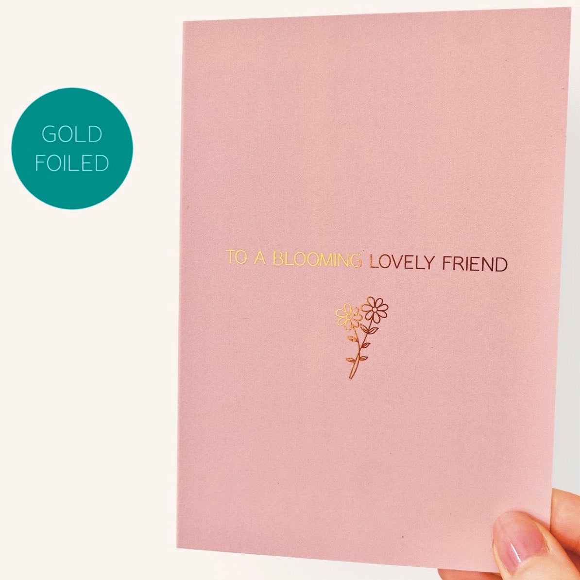 Pink card with gold foiled text 'To a blooming lovely friend' and a flower design, held by a hand.