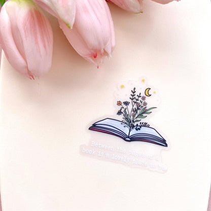 Decorative clear sticker with an open book design and floral elements and a 'Between the pages of a book is a lovely place to be' quote against a cream background