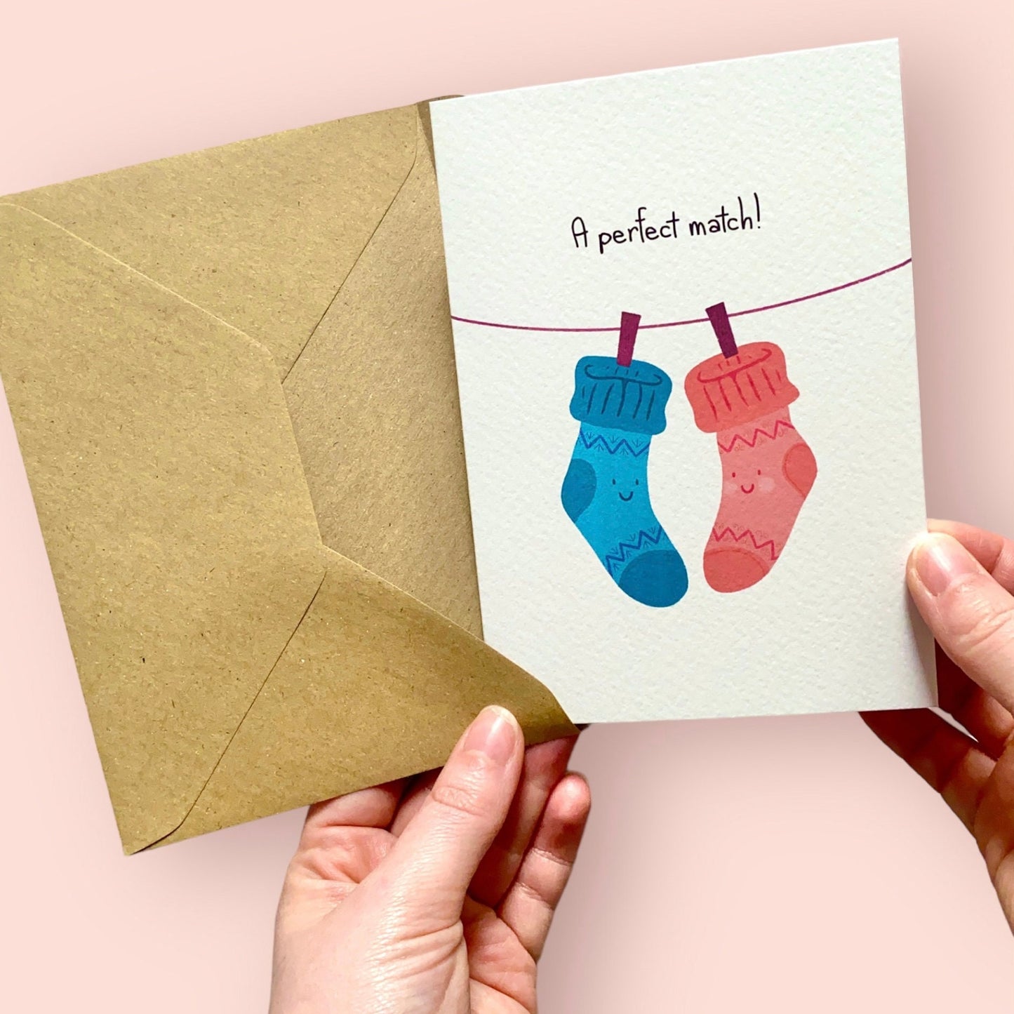 Greeting card with two socks on a line and 'A perfect match!' text, held by hands with a pink background.
