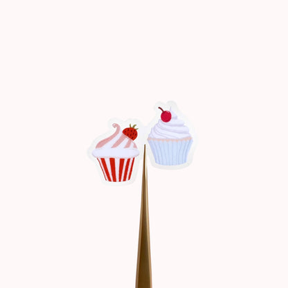 A sticker with two tiny cupcakes in pink and blue against a cream background.