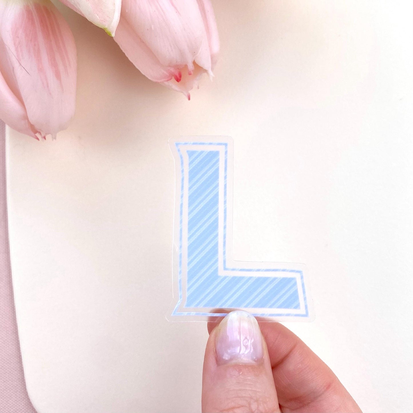 Hand holding a blue letter 'L' sticker with pink tulips in the background